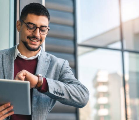 Young happy businessman looking at watch while holding digital tablet outside of modern building on a sunny day. Male CEO checking time for his next appointment.